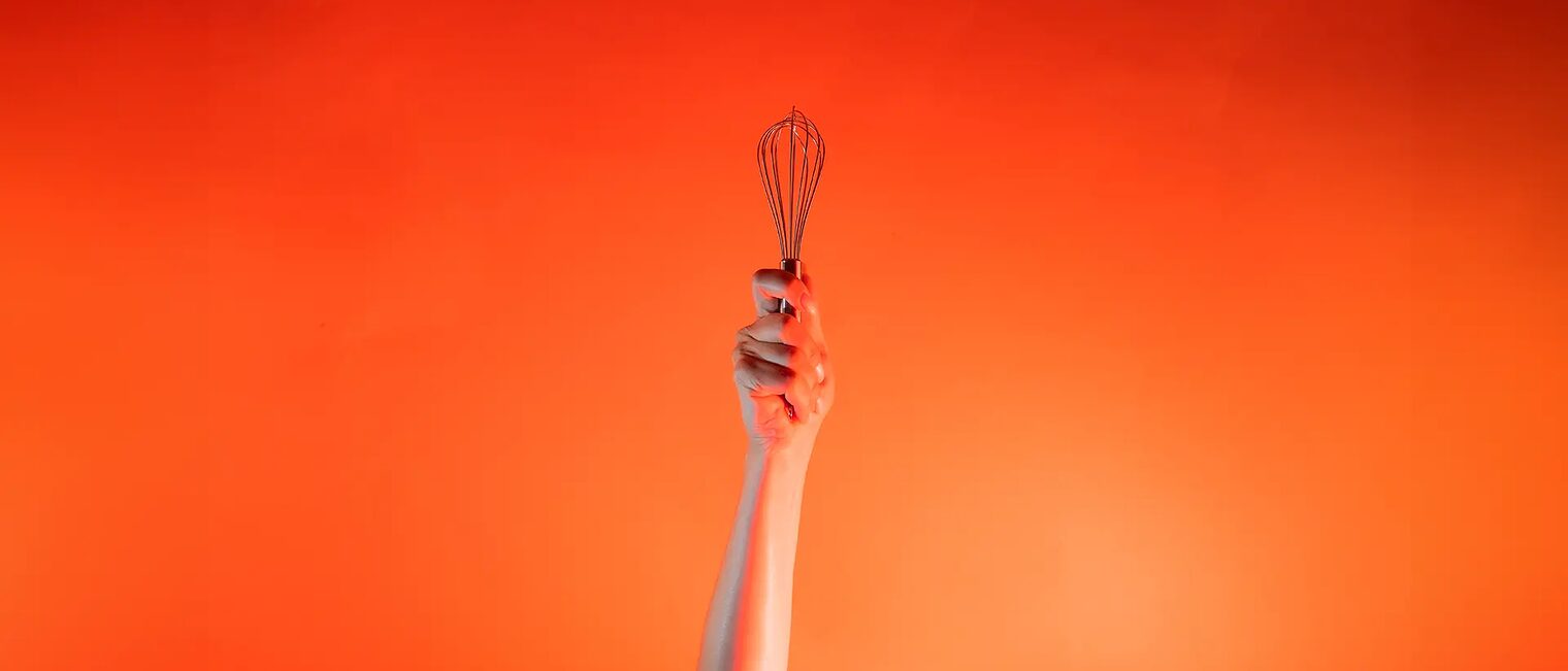 Ein Arm ist vor orangenem Hintergrund senkrecht nach oben gestreckt und in der Hand wird ein Schneebesen gehalten.