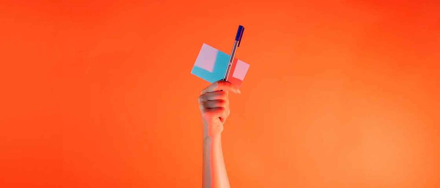 Ein Arm ist vor orangenem Hintergrund senkrecht nach oben gestreckt und hält in der Hand einen Notizblock und eine Stift.