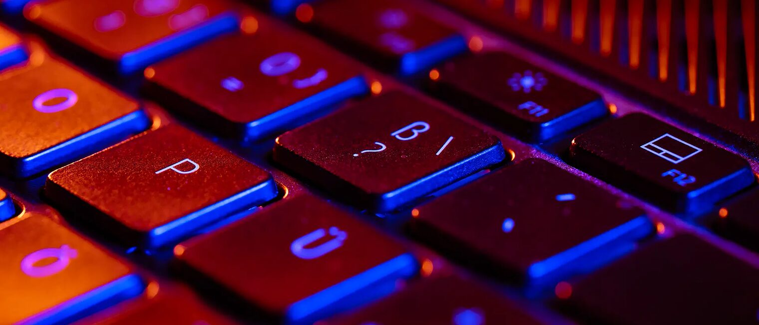 Fragezeichentaste für FAQs Die Tastatur eines Laptops wird orange und blau angeleuchtet. Im Zentrum und Focus des Fotos: Die Fragezeichentaste, als Symbol für die FAQs.